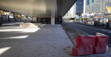 Las obras de mejora de accesibilidad bajo el puente de Raimundo Fernández Villaverde en Madrid han entrado en su fase final, y se espera que estén concluidas a principios de 2025. Este proyecto, liderado por el Ayuntamiento de Madrid, tiene como objetivo transformar el espacio, facilitando su uso peatonal, garantizando la accesibilidad universal y poniendo en valor el grupo escultórico existente. Con una inversión de 600.000 euros y una superficie de actuación de 2.937 metros cuadrados, esta intervención busca reconfigurar un área clave de la ciudad, combinando funcionalidad, estética y sostenibilidad. Una intervención integral para los peatones El corazón del proyecto reside en la creación de un plano continuo de suelo, eliminando escalonamientos para asegurar un tránsito más cómodo y accesible. Esta medida permite a las personas con movilidad reducida desplazarse sin barreras físicas, cumpliendo con los estándares de accesibilidad universal. Además, el diseño incluye la instalación de zonas de asiento, compuestas por bancos y cubos, que invitan a los transeúntes a hacer una pausa en su camino. La mejora no solo es funcional, sino también estética. Los pilares y el techo del puente se han revestido con vinilos ilustrados con imágenes de pinos, un guiño al entorno natural de una calle que se distingue por contar con esta especie arbórea. Este toque visual transforma un espacio antes desaprovechado en un lugar más acogedor y atractivo. Zonas verdes que enriquecen el entorno Un elemento destacado de la remodelación es la incorporación de bandas ajardinadas laterales, que no solo embellecen el espacio, sino que también contribuyen a mejorar la calidad ambiental de la zona. La elección de la vegetación ha sido cuidadosa, respetando las recomendaciones de la Ordenanza de Gestión y Uso Eficiente del Agua en Madrid. Las gramíneas, plantas adaptadas al clima local y con bajos requerimientos de mantenimiento y agua, serán las protagonistas de estas áreas verdes. Esta combinación de diseño funcional y elementos naturales refuerza el compromiso del Ayuntamiento con un urbanismo sostenible, equilibrando las necesidades de los peatones con la mejora del paisaje urbano. Materiales innovadores y sostenibles La pavimentación del área se realiza con hormigón continuo antideslizante, garantizando la seguridad de los peatones, especialmente en condiciones de lluvia. En las bandas laterales no ajardinadas, se ha optado por adoquines de granito, un material duradero que aporta un acabado elegante. Uno de los cambios más llamativos es la sustitución del antiguo estanque, que se encontraba vacío y deteriorado. Este ha sido reemplazado por un pavimento de acero inoxidable con acabado de brillo espejo, que recrea el efecto de reflejo del agua y aporta un toque moderno al diseño. Para delimitar los distintos pavimentos, se utilizan pletinas de acero galvanizado, mientras que el perímetro del área está definido por bordillos de hormigón. Una apuesta por la accesibilidad universal La delegada de Obras y Equipamientos, Paloma García Romero, visitó recientemente las obras junto a los concejales de Chamartín, Yolanda Estrada, y de Chamberí, Jaime González Taboada. Durante su recorrido, destacaron la importancia de este proyecto como parte de un compromiso más amplio del Ayuntamiento para hacer de Madrid una ciudad más accesible y sostenible. “La transformación de este espacio no solo mejora la movilidad de los peatones, sino que también pone en valor un entorno que estaba infrautilizado. Es un ejemplo de cómo los espacios públicos pueden ser accesibles, funcionales y estéticamente agradables al mismo tiempo”, señaló García Romero. Una nueva vida para un espacio olvidado Bajo el puente de Raimundo Fernández Villaverde, este proyecto representa mucho más que una simple mejora estructural. Es una reinvención del espacio público, que lo convierte en un lugar más habitable y atractivo tanto para los vecinos como para los visitantes. La combinación de diseño inclusivo, sostenibilidad ambiental y materiales innovadores refleja un enfoque contemporáneo del urbanismo que prioriza las necesidades de las personas. Hacia un Madrid más accesible y sostenible Con la finalización prevista de las obras en 2025, el puente de Raimundo Fernández Villaverde se unirá a una lista creciente de proyectos que buscan hacer de Madrid una ciudad más accesible, habitable y comprometida con el bienestar de sus ciudadanos. Este espacio renovado promete ser no solo un paso más seguro para los peatones, sino también un ejemplo de cómo las ciudades pueden integrar diseño, funcionalidad y sostenibilidad para mejorar la calidad de vida urbana.